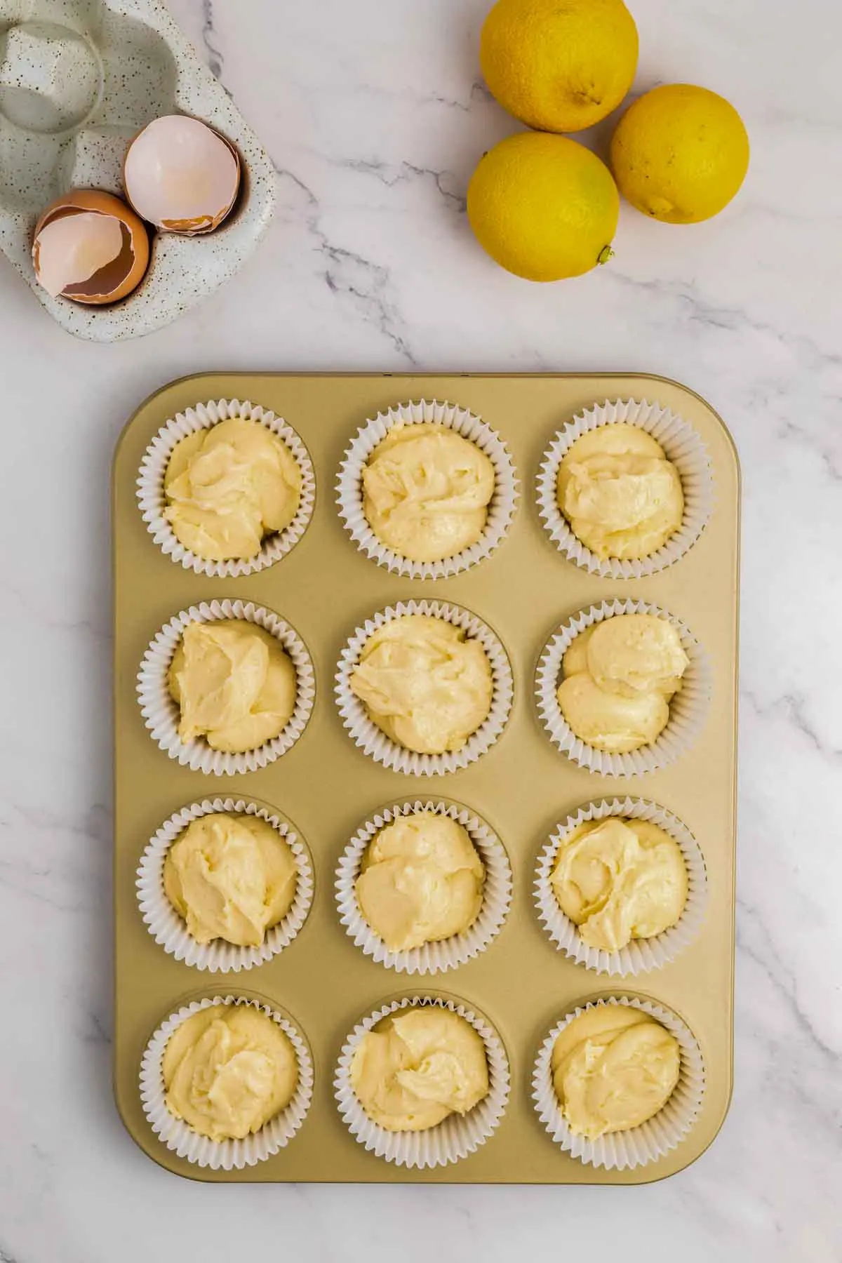 lemon cupcake batter in a muffin tin on counter with lemons and empty egg shells above