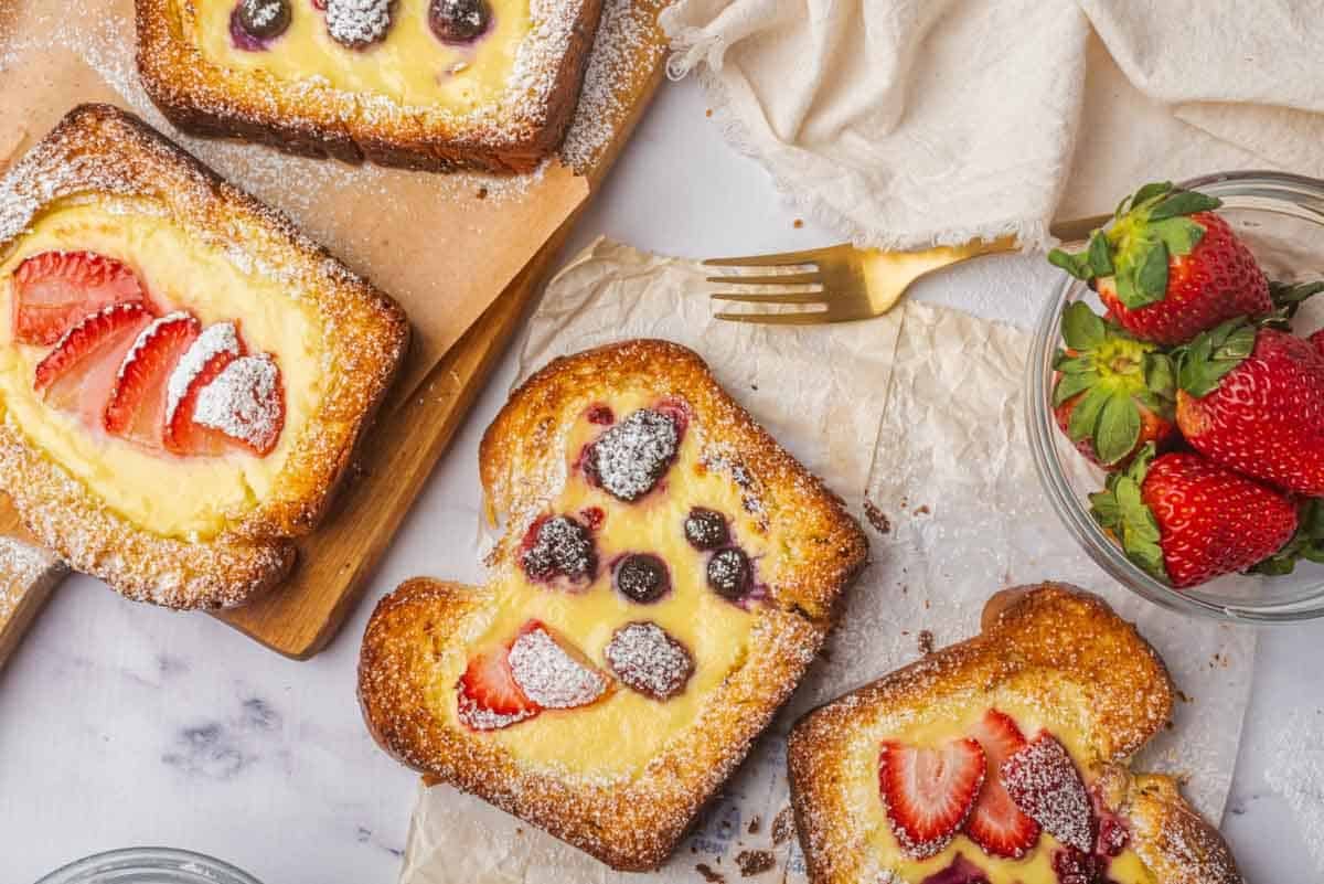 a few pieces of toast filled with yogurt custard topped with fruit, with a fork and bowl strawberries