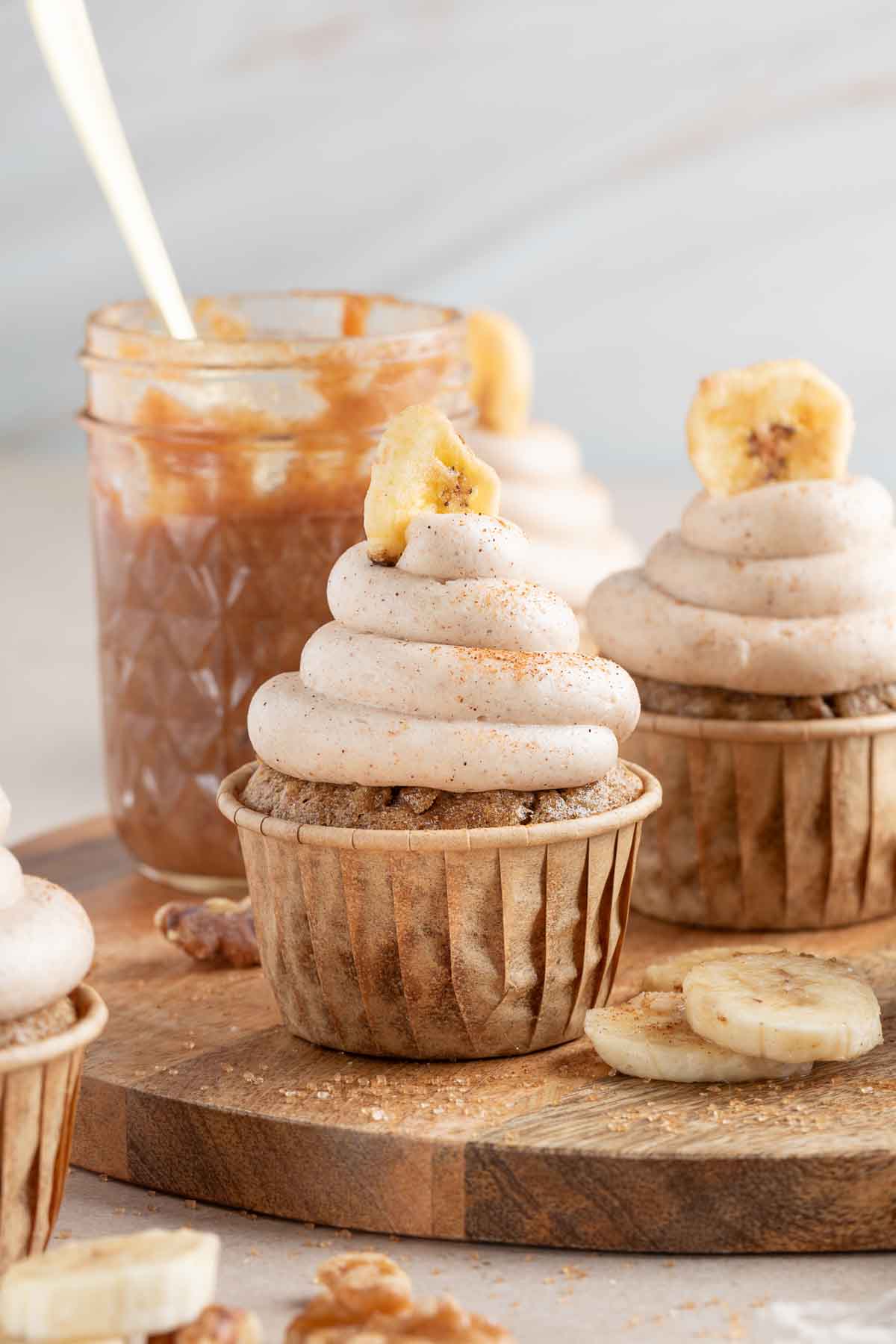 banana bread cupcakes in liners topped with cream cheese frosting and a dried banana chip with the jar of banana jam in background