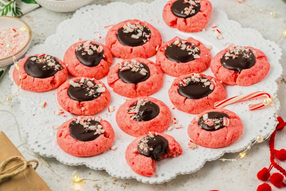 13 peppermint thumbprint cookies with chocolate ganache on a white plate with one cookie with a bite out of it