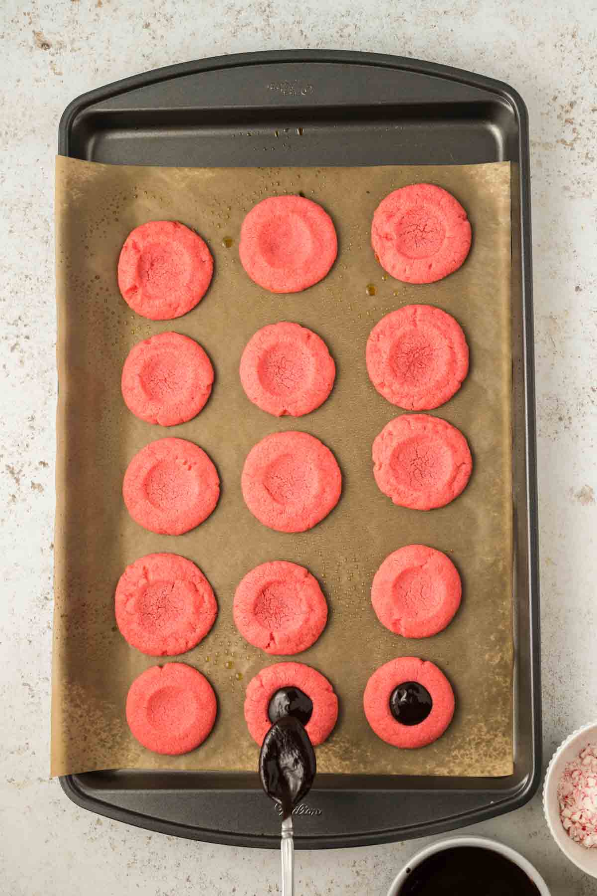 a dozen peppermint cookie dough circles on a baking sheet with 2 cookies filled with chocolate ganache by spoon