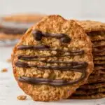 a stack of oatmeal lace cookies with one cookie leaning next to it with chocolate drizzle