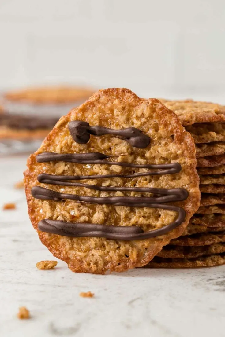 a stack of oatmeal lace cookies with one cookie leaning next to it with chocolate drizzle