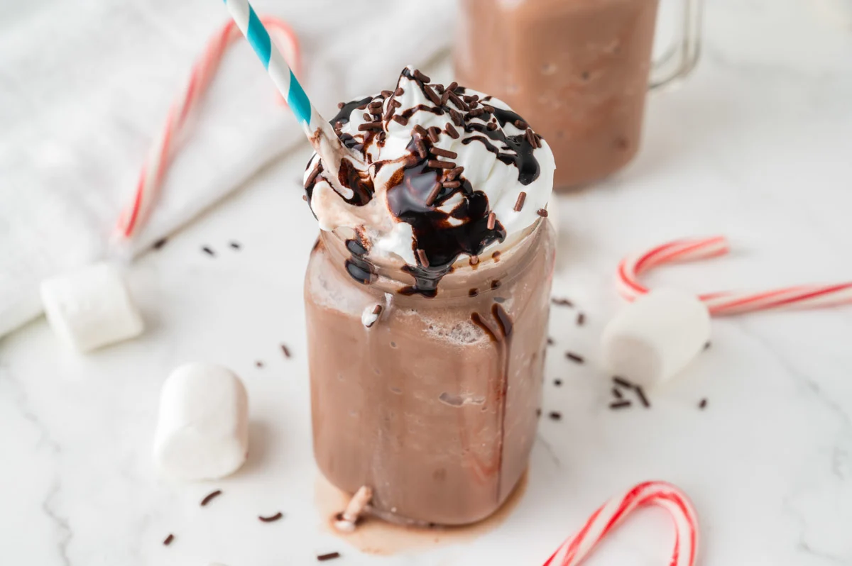 Frozen Hot Cocoa in a mason jar topped with whipped cream, chocolate syrup and chocolate sprinkles with a blue and white straw, with candy canes and marshmallows around