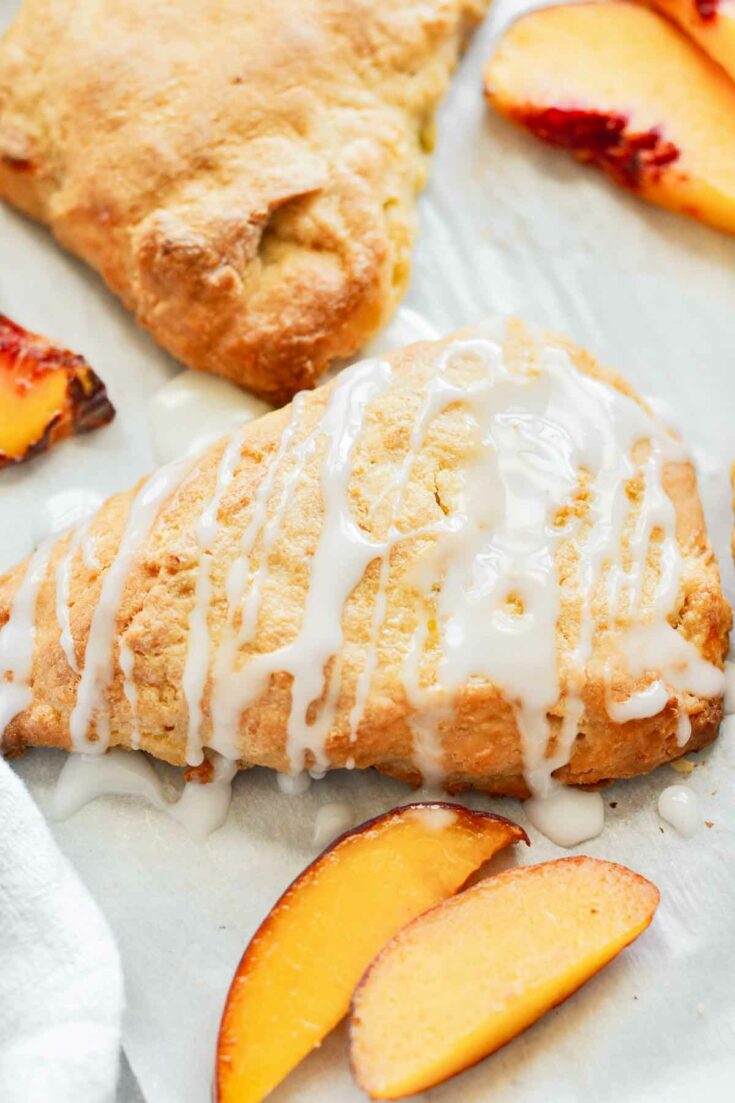 a peach scone with drizzled glaze with a few fresh peach slices laying next to it