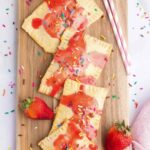 4 homemade strawberry pop tarts of wooden cutting board with a few fresh strawberries around it and pink straws