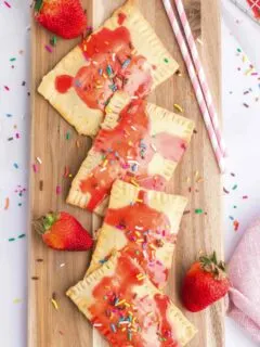 4 homemade strawberry pop tarts of wooden cutting board with a few fresh strawberries around it and pink straws