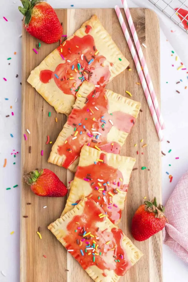 4 homemade strawberry pop tarts of wooden cutting board with a few fresh strawberries around it and pink straws