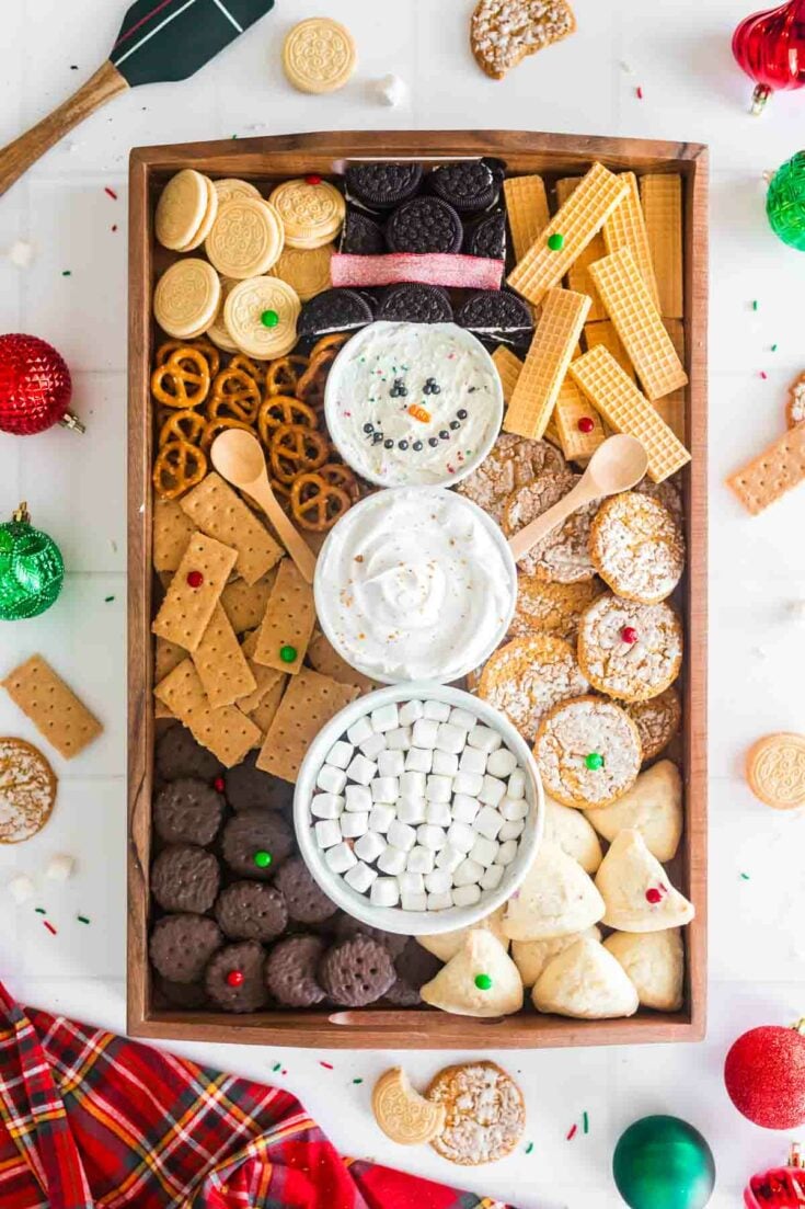 Frosty snowman dessert dip board with cookies and dippers surrounding it on a wooden tray