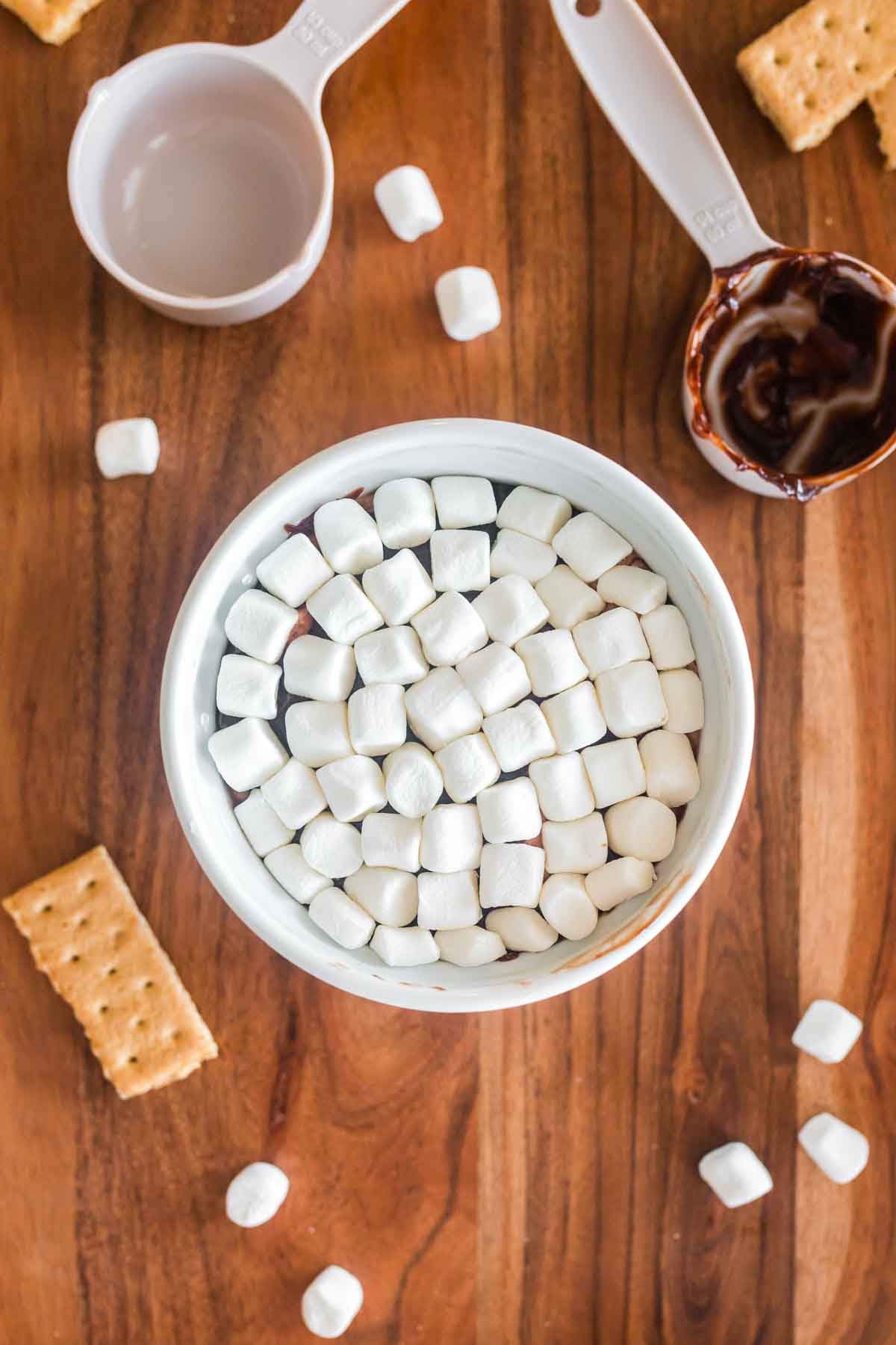 hot chocolate dip in white ramekin covered in mini marshmallows, with a small graham cracker , extra mini marshmallows and hot fudge cup on side, resting on wood background