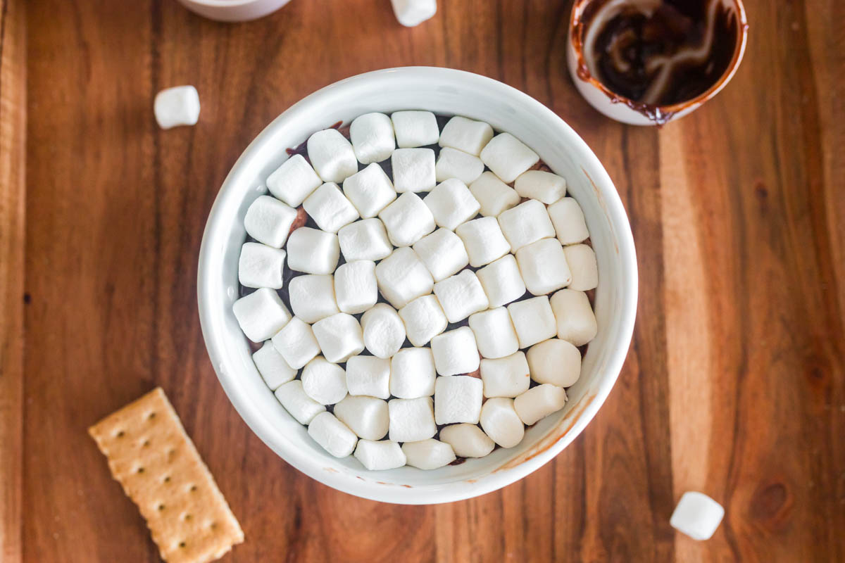 hot chocolate dip in white ramekin covered in mini marshmallows, with a small graham cracker and hot fudge cup on side, resting on wood background