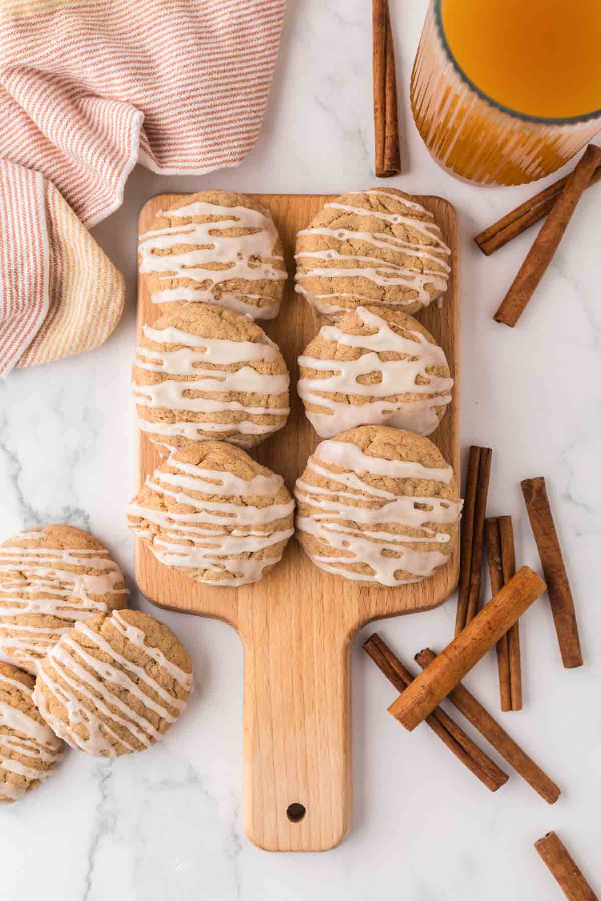 6 apple cider cookies on wooden serving tray, with cinnamon sticks and glass of apple cider with pink napkin.