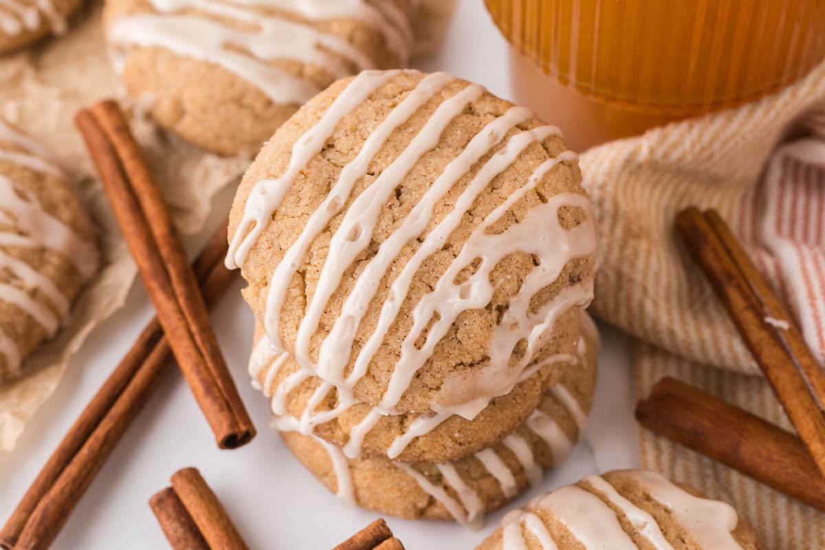 stacked iced apple cider cookies with cinnamon sticks and a glass of apple cider.