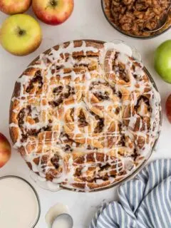 baked apple pie cinnamon rolls in baking dish drizzled with icing, with apples and ingredients next to it on the counter.