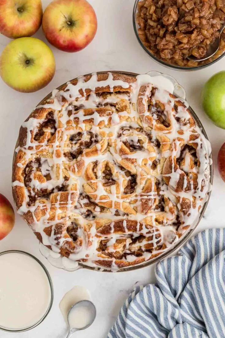 baked apple pie cinnamon rolls in baking dish drizzled with icing, with apples and ingredients next to it on the counter.