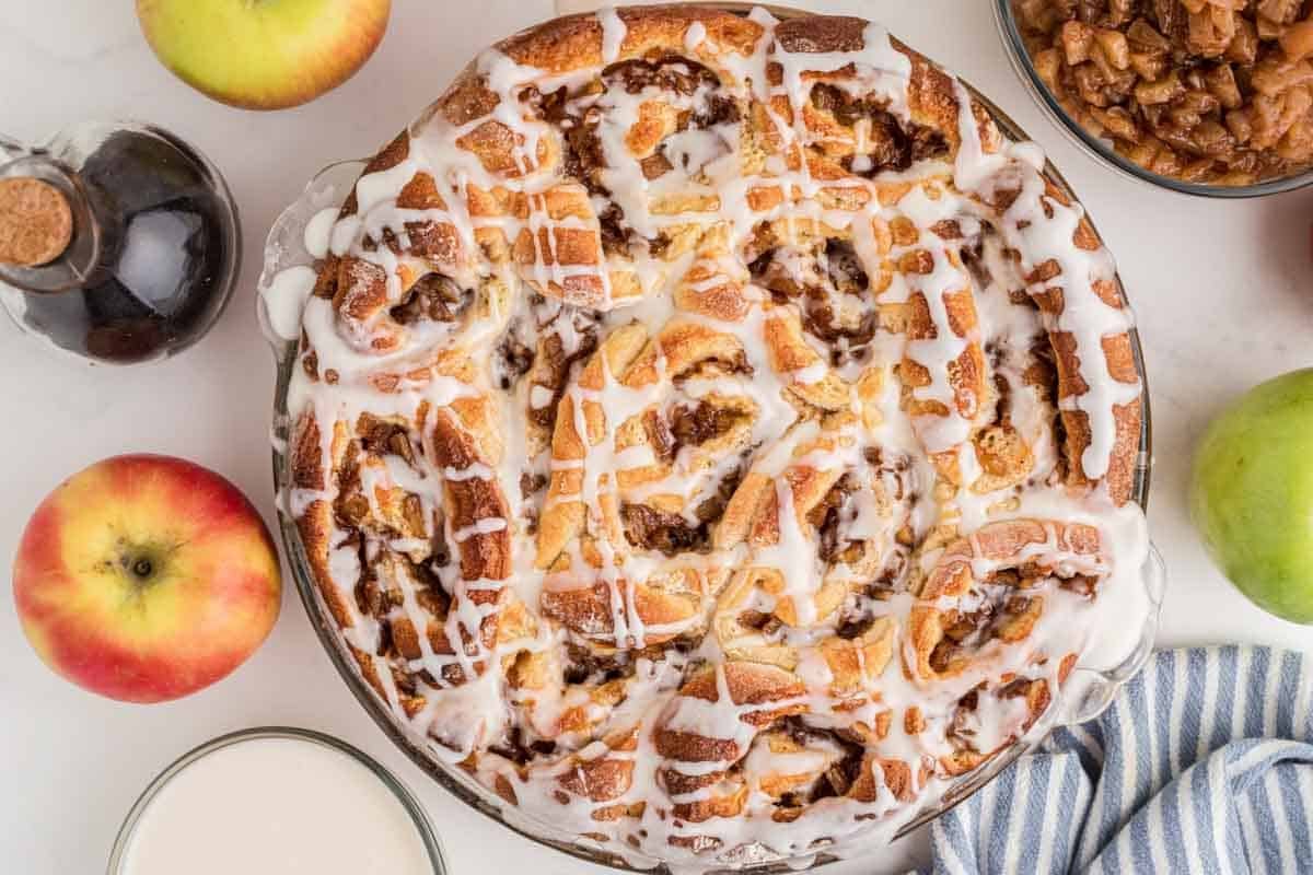 baked apple pie cinnamon rolls in baking dish drizzled with icing, with apples and ingredients next to it on the counter.