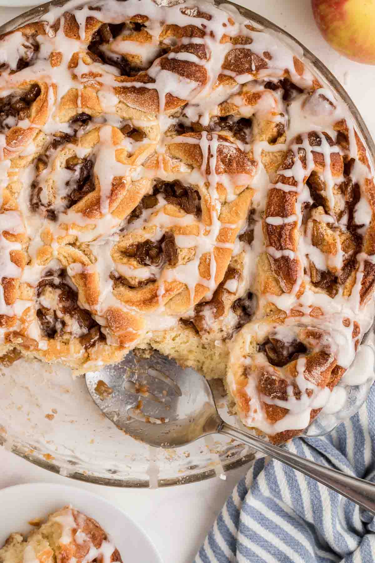 baked apple pie filled cinnamon rolls with frosting in glass baking dish, with a couple rolls taken out.