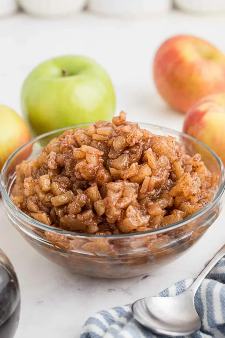 Clear mixing bowl of homemade apple pie filling with whole apples in background.