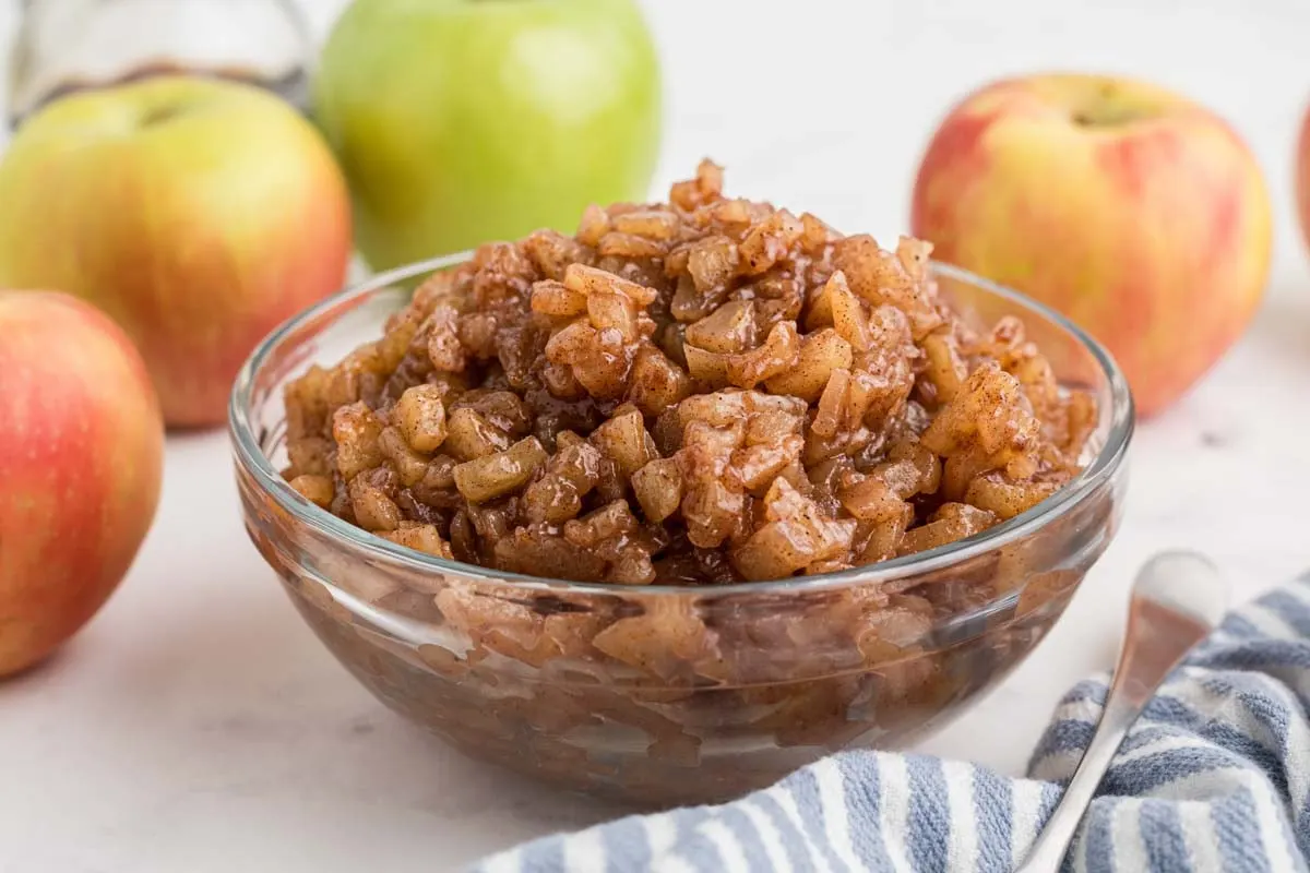 Clear mixing bowl of homemade apple pie filling with whole apples next to it.