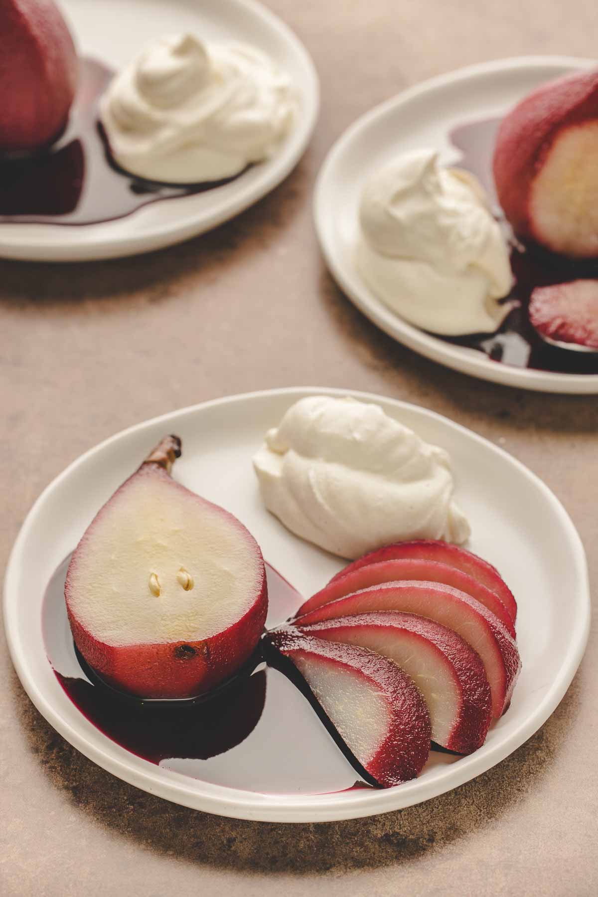 half a red wine poached pear with extra pear slices on white plate, whipped mascarpone cream next to it.