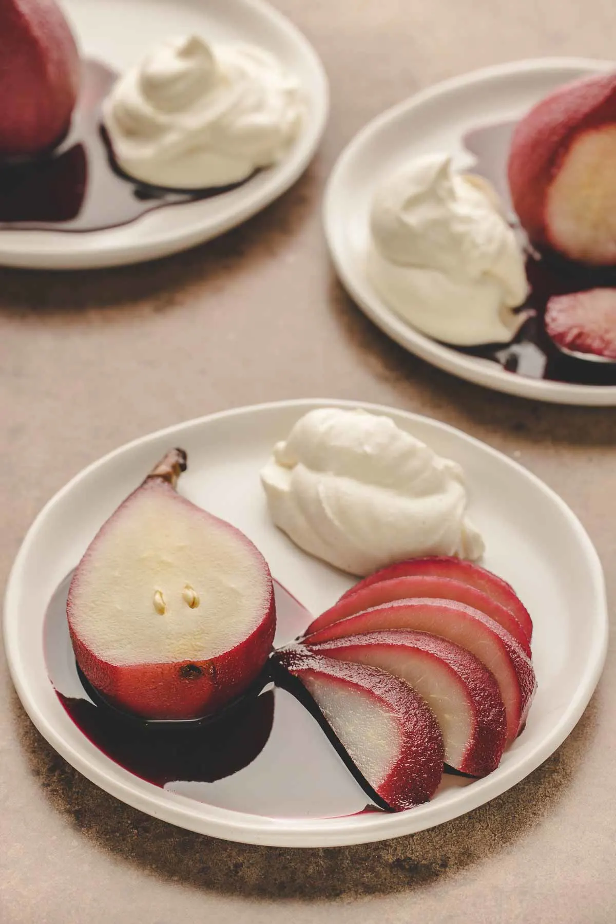 half a red wine poached pear with extra pear slices on white plate, whipped mascarpone cream next to it.