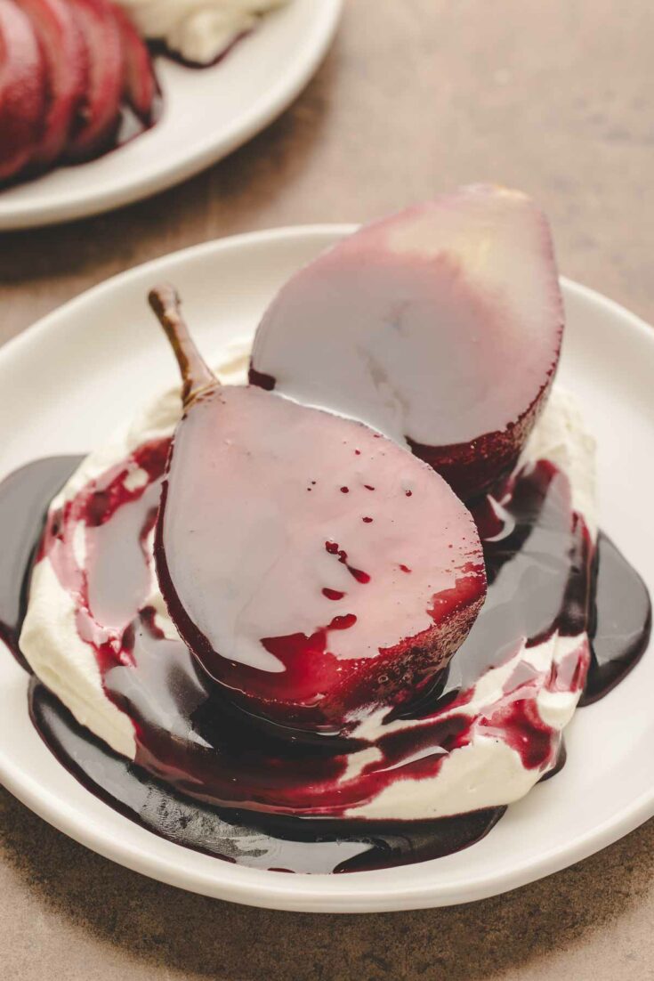 2 halves of red wine poached pear sitting on vanilla whipped mascarpone cream and extra red wine reduction sauce on white plate