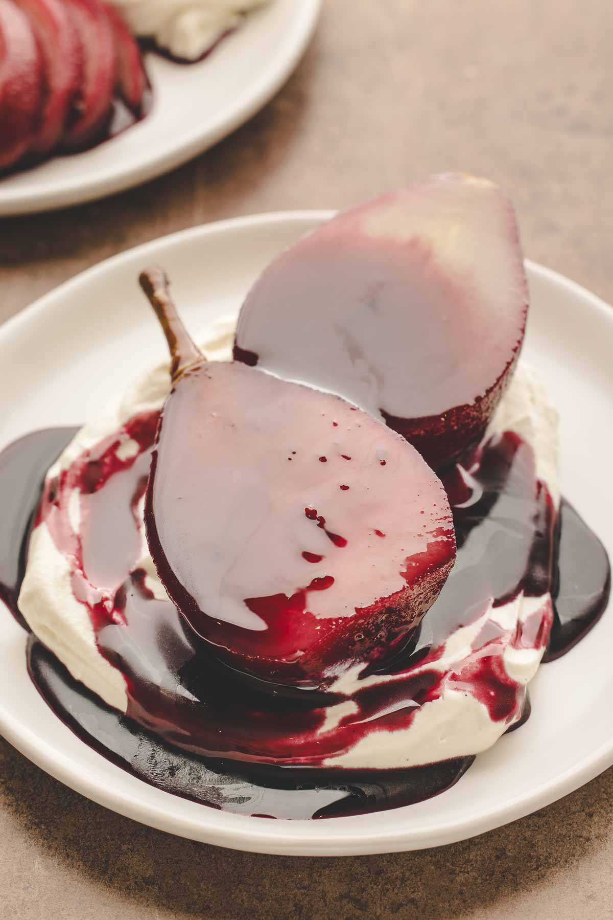 2 halves of red wine poached pear sitting on vanilla whipped mascarpone cream and extra red wine reduction sauce on white plate. 