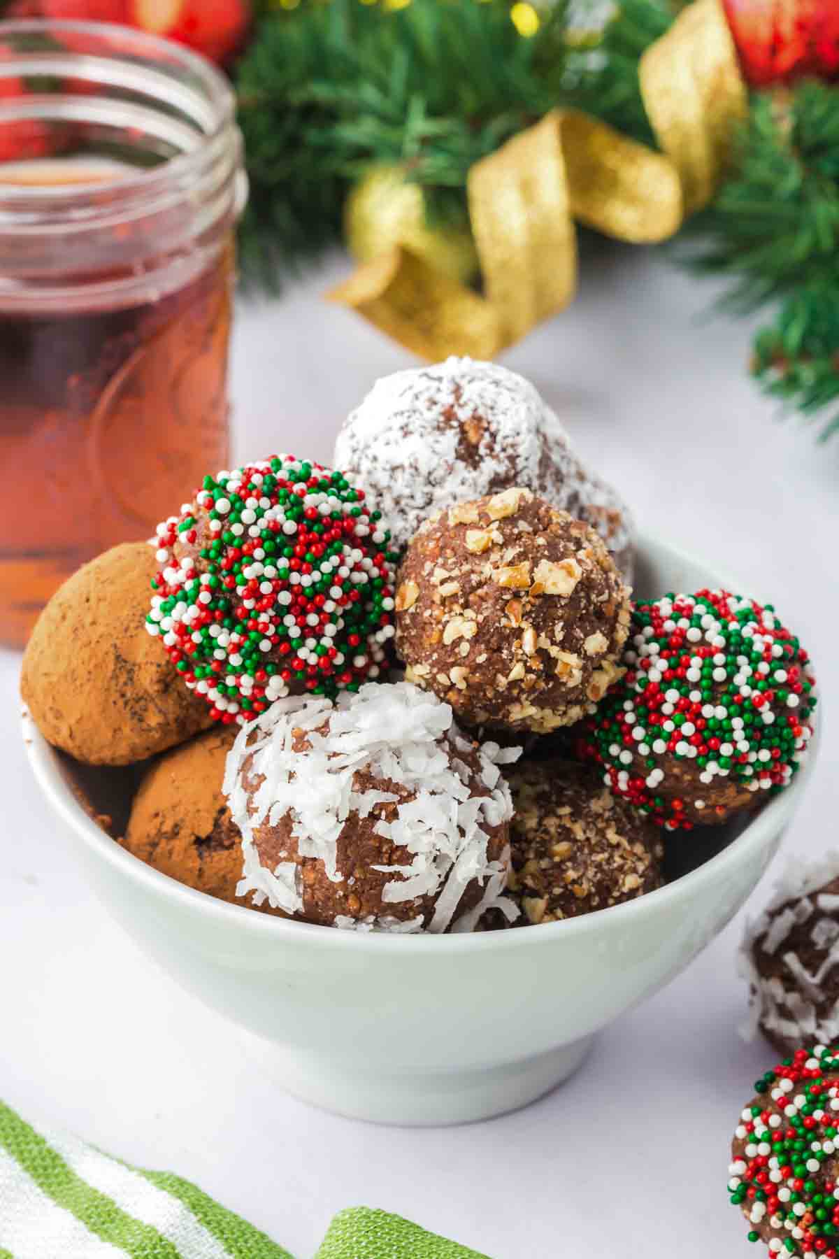 Chocolate Rum Balls with different toppings in a white bowl and Christmas decorations, a glass of rum in the background.